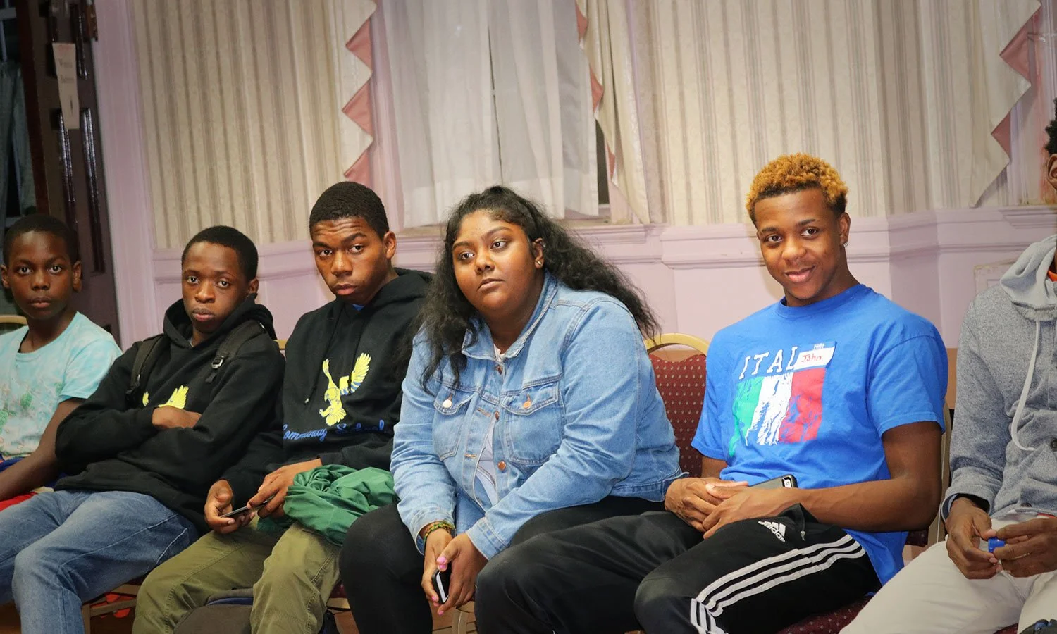 Group of young people sitting and listening in a room with pink walls and curtains.