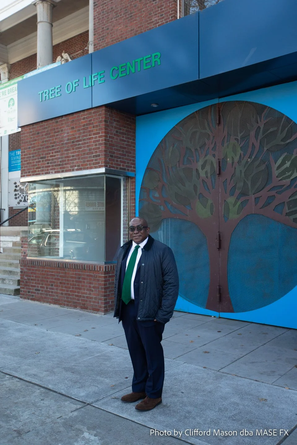 A man dressed in a suit and tie, wearing sunglasses, standing outside the Tree of Life Center, a building with a blue and brick exterior and a large tree-themed artwork on the door.