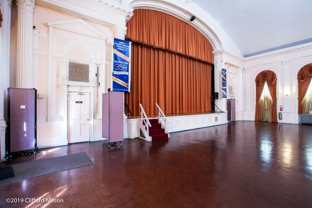 Empty stage with closed orange curtains in a decorated hall with white paneled walls and tall windows with matching orange drapes.