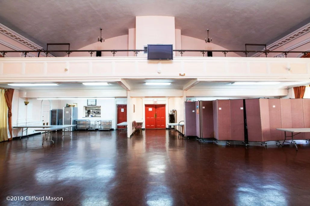 Empty banquet hall or event space with a polished wooden floor, a loft balcony with black railing, and old-fashioned chandeliers. There are tables and divider screens along the walls, and a small kitchenette area in the background.