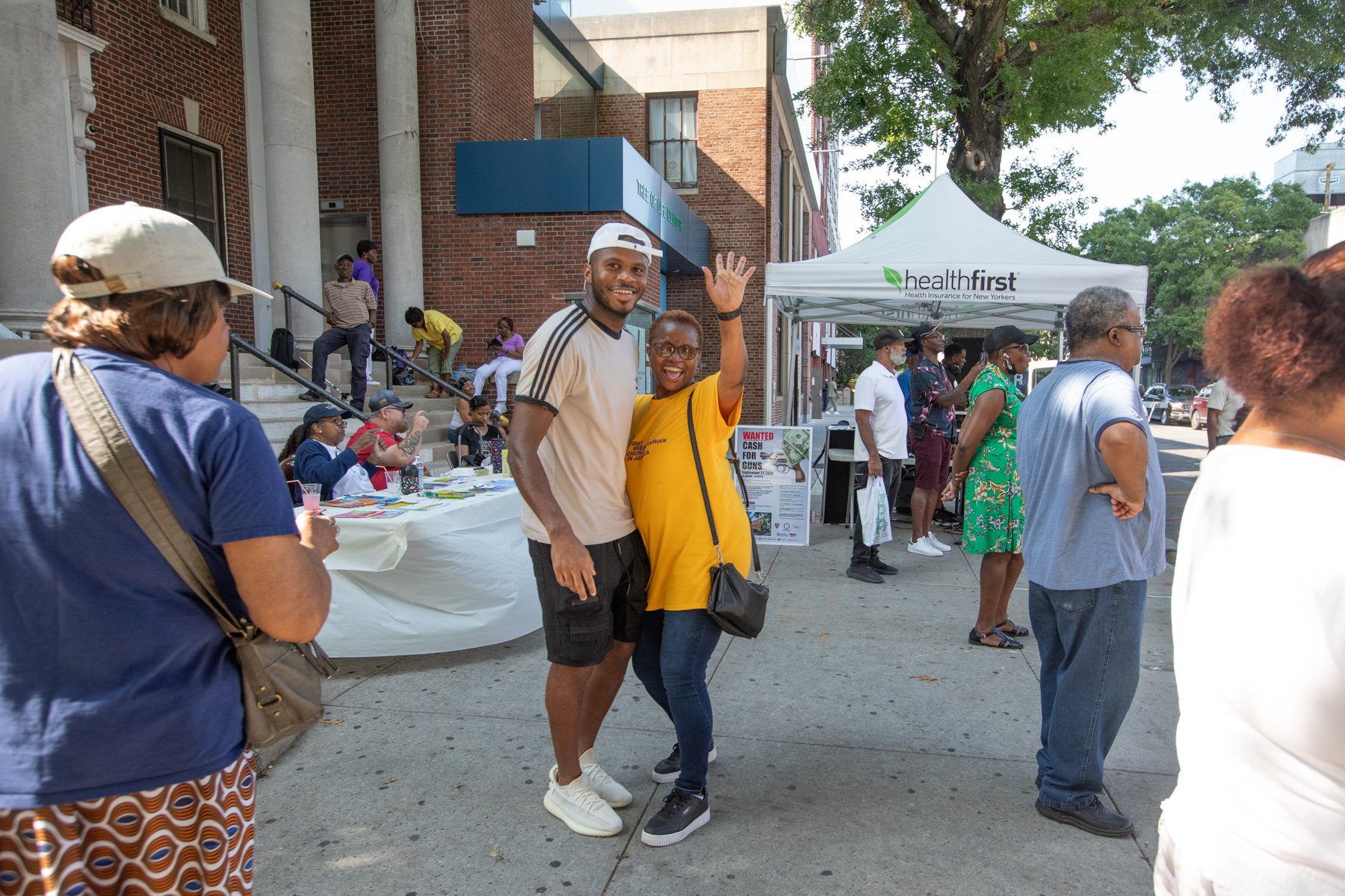 People attending an outdoor community event on a city street, with a white tent displaying 'health first' logo, a table with flyers, and a group of diverse individuals socializing and waving.
