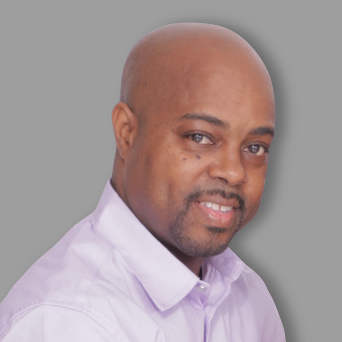 A portrait of a man with a shaved head, wearing a light purple collared shirt, smiling slightly against a plain gray background.