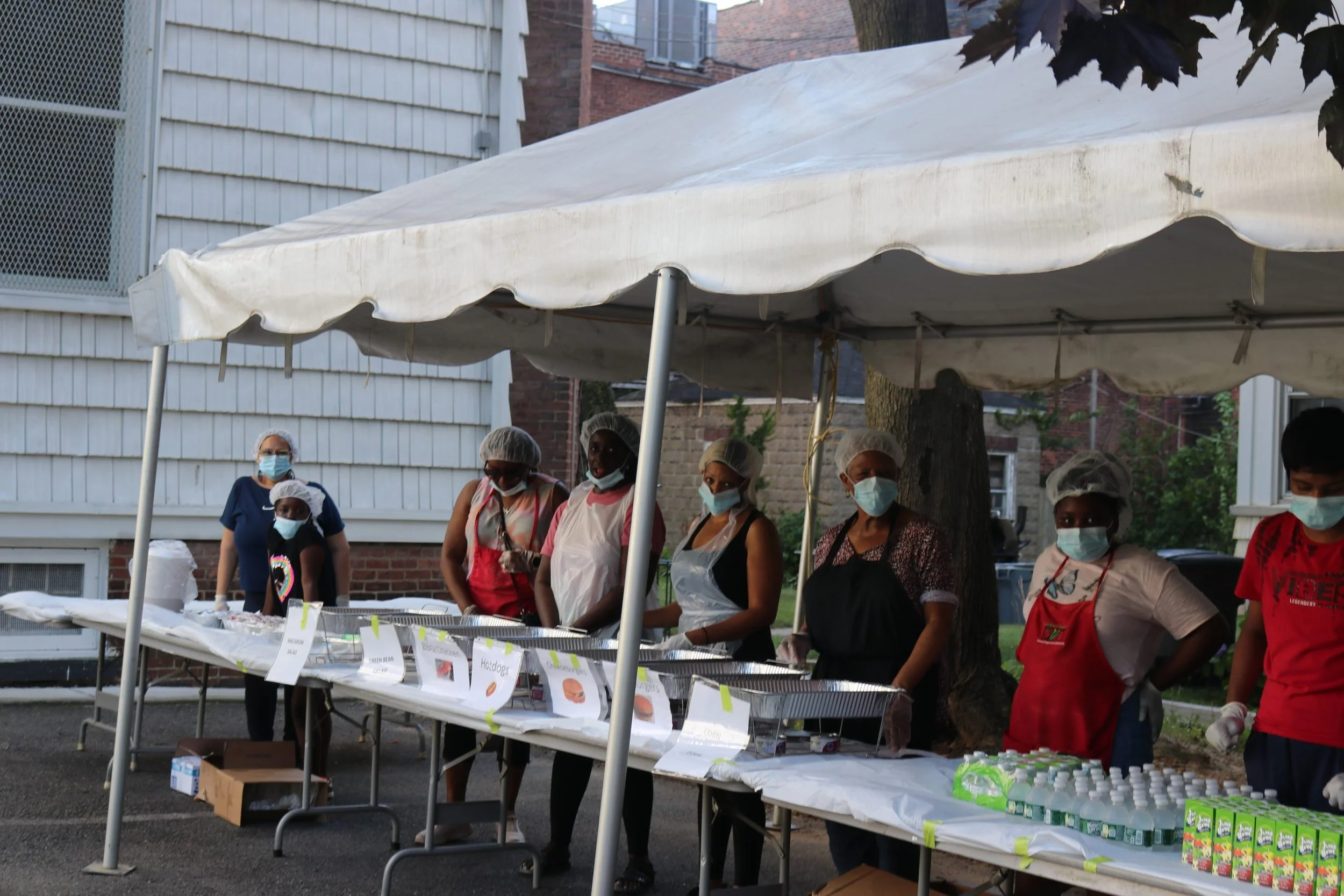 People wearing masks and aprons standing behind an outdoor food and drink stand with labeled trays of hot dogs, snacks, and bottled drinks. They appear to be preparing and serving food at an event.