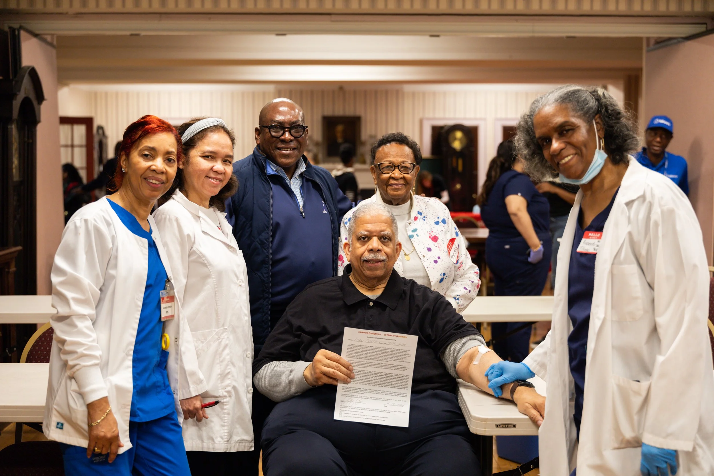 Group of six diverse healthcare professionals, including nurses and a doctor, posing together at a blood donation event. One person is seated, holding a donation form and has an arm exposed for blood donation. The group is smiling in a well-lit, indoor setting.