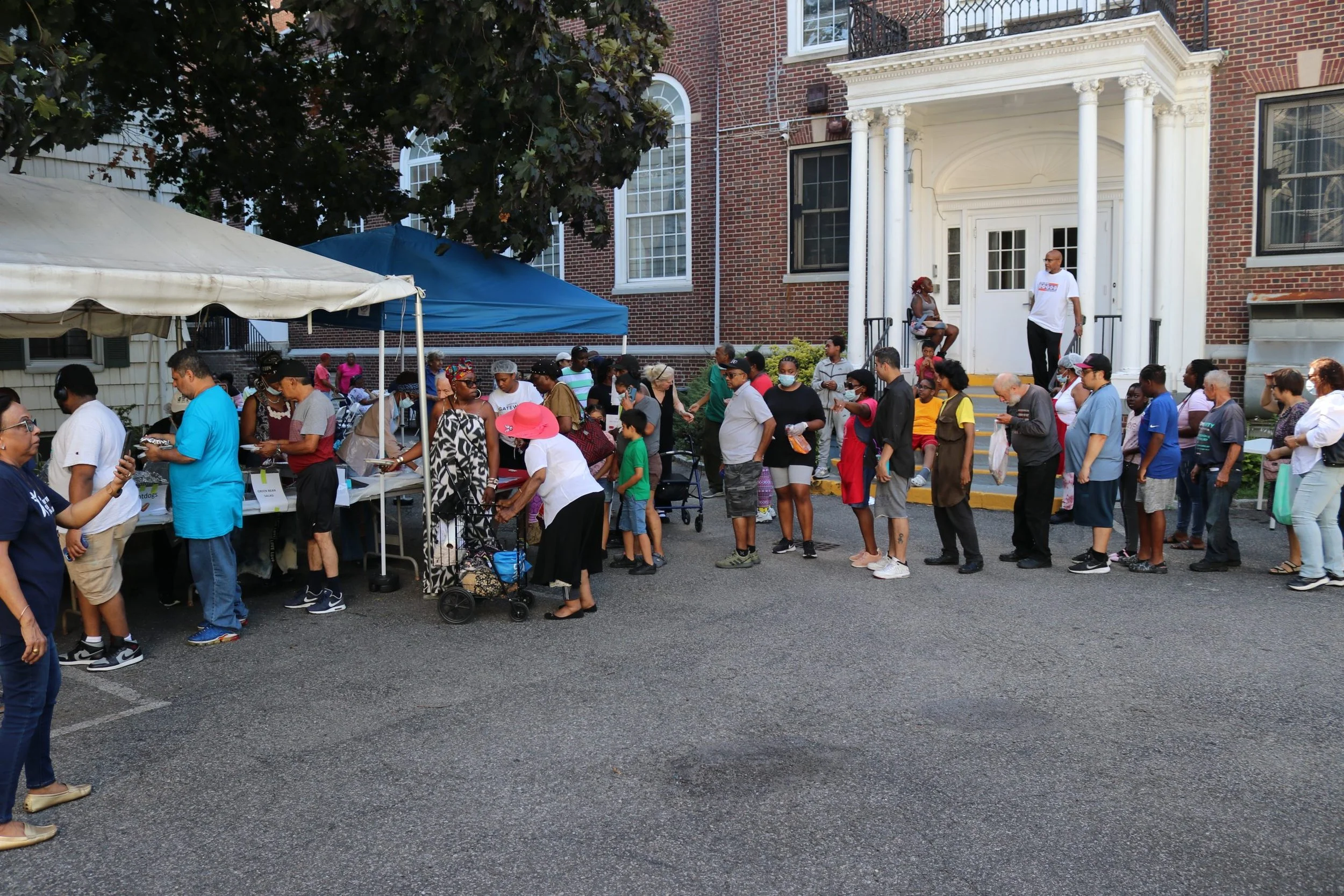 People standing in a line outside a building with a brick facade and white columns, possibly waiting for a community event or food distribution under tents.