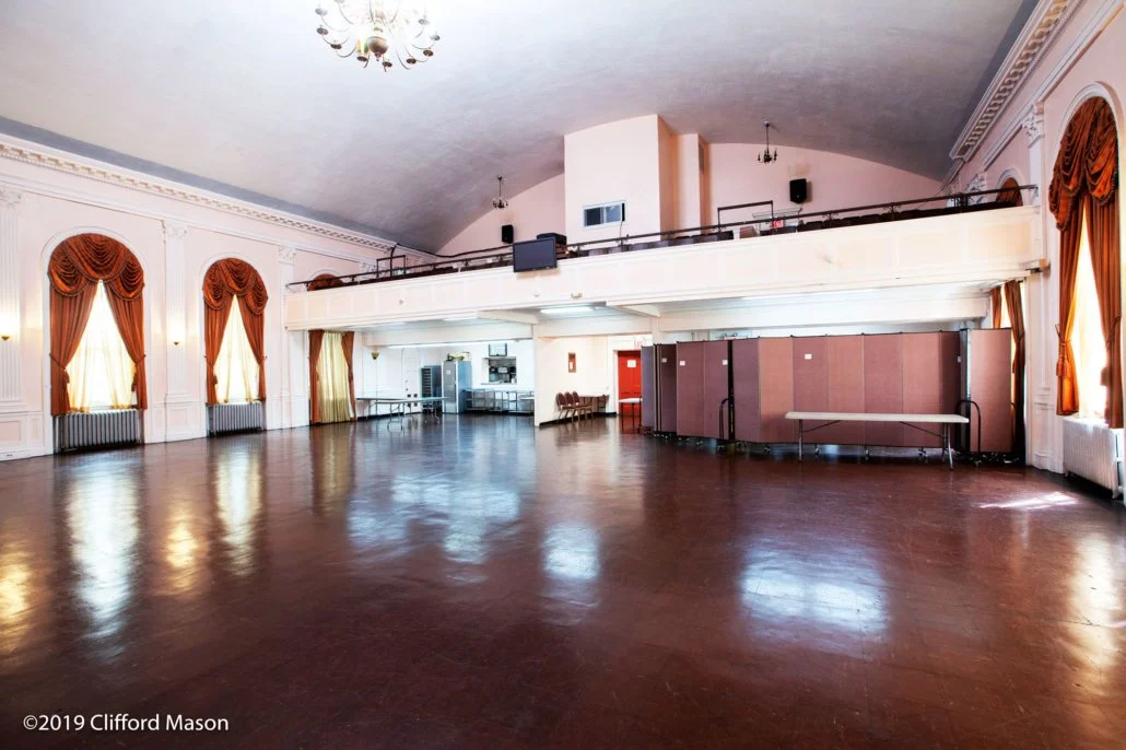 Large, empty ballroom with polished wooden floor, tall windows with gold curtains, and elegant white moldings. There's a balcony with a railing, a few tables, and equipment in the background.