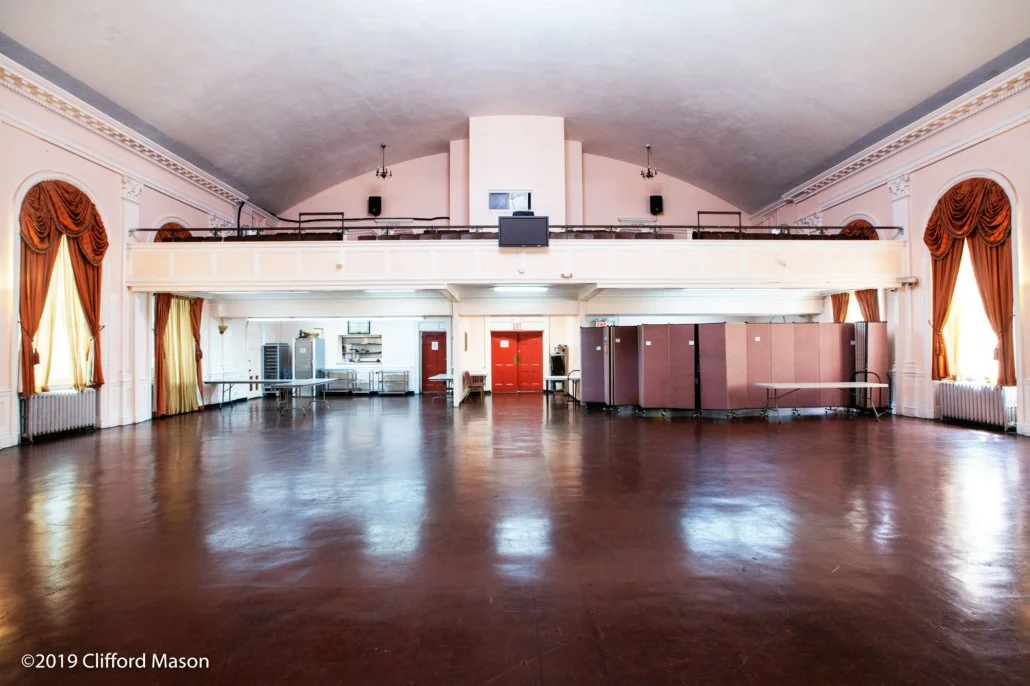 Empty banquet hall with wooden floors, large windows with gold curtains, and a raised balcony with black railings. There are a few tables and equipment along the back wall, and pink folding partitions on the right. The room is well-lit with natural l