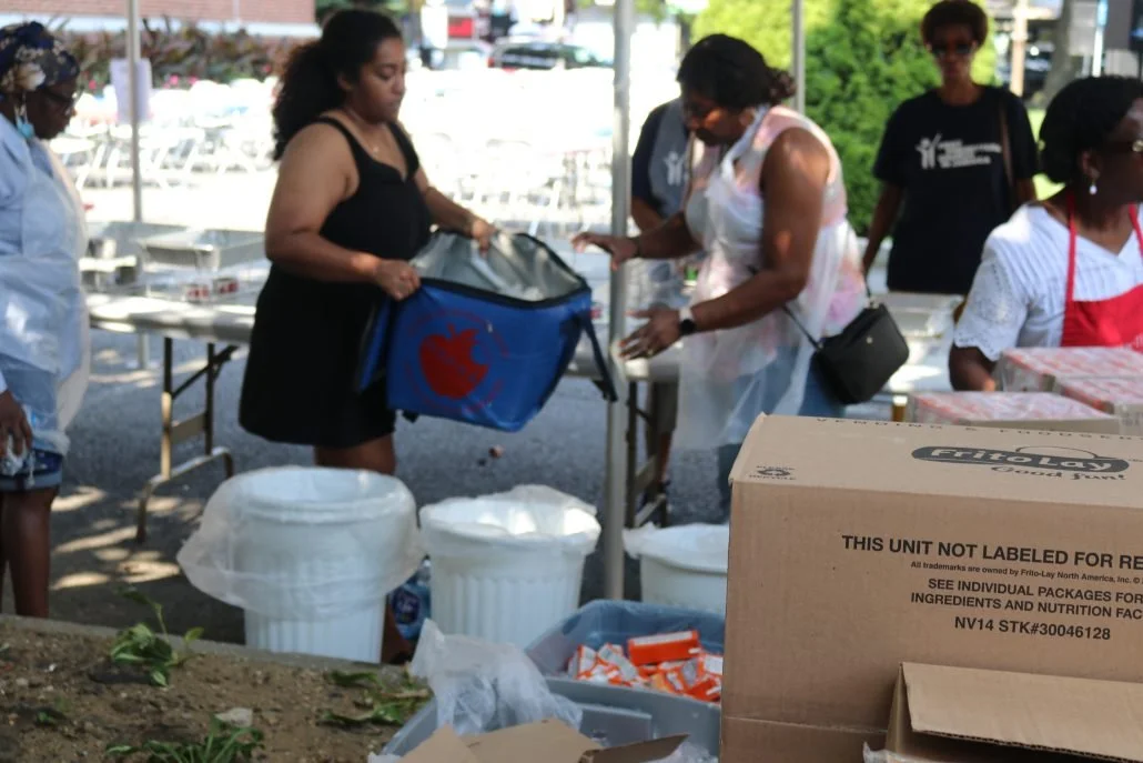 Group of women at an outdoor event, some carrying packages and standing near tables with boxes and earthware containers, under a canopy with trees in the background.