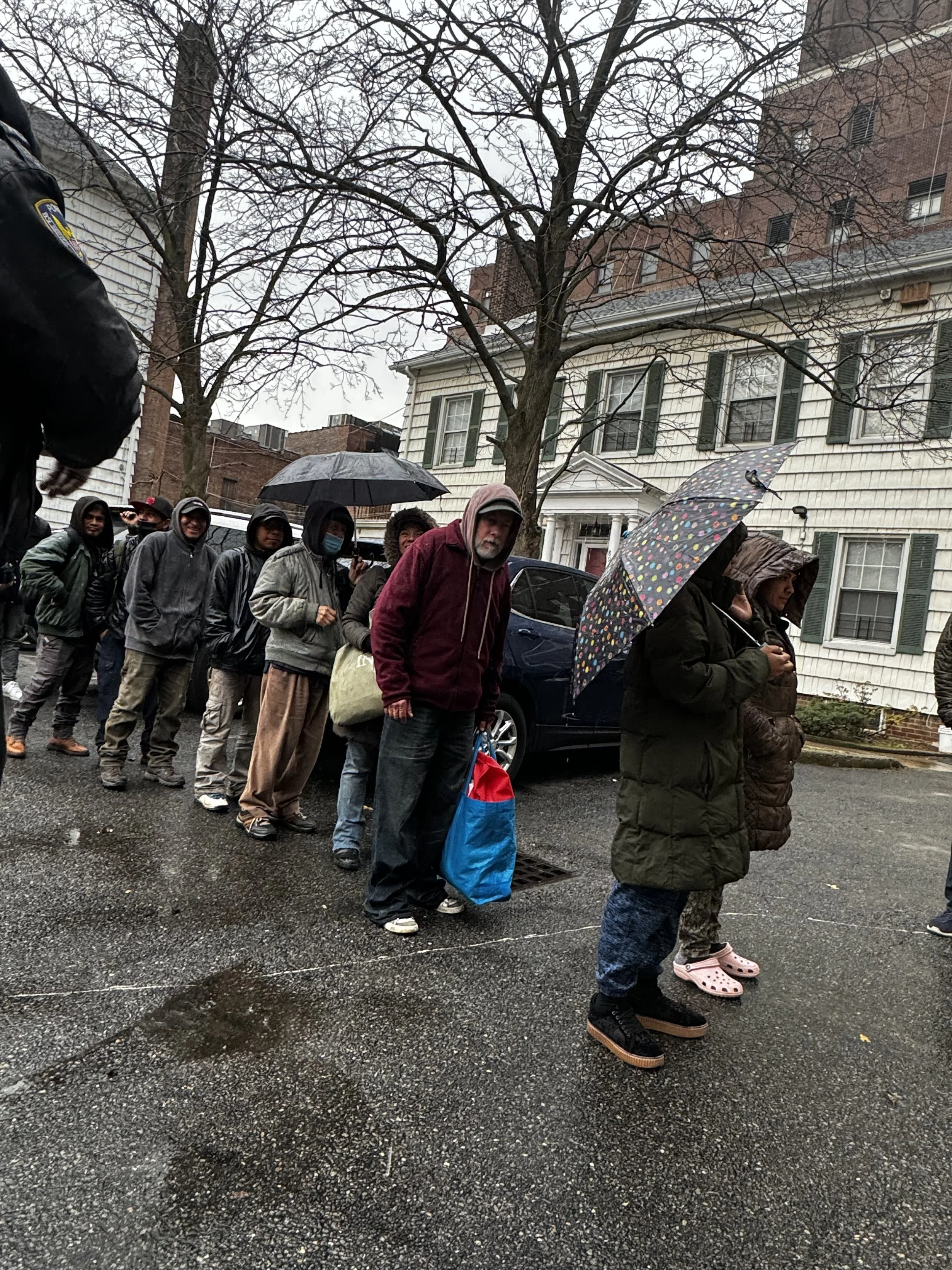 A line of people standing outdoors on a rainy day, holding umbrellas, wearing warm clothing, with trees and residential houses in the background.
