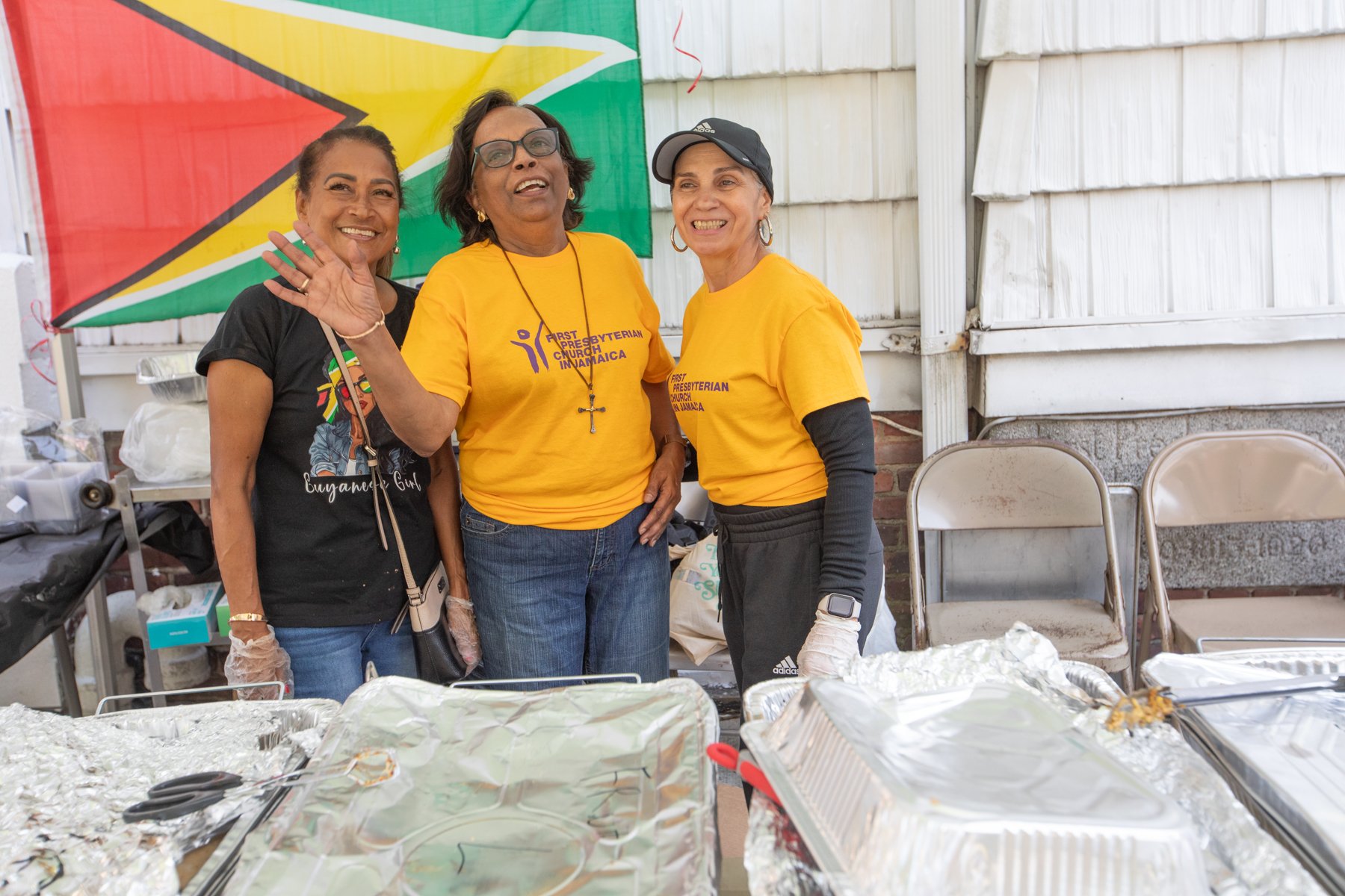 Three women smiling and posing together at a community event, with trays of food or supplies in front of them and a banner with the flag of Guyana in the background.