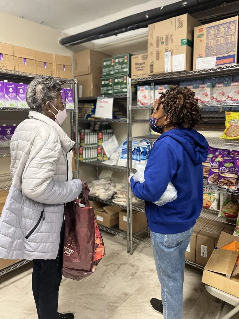 Two women wearing masks and winter clothing standing in a grocery store aisle, engaging in conversation. One woman is holding a shopping bag, and the shelves are stocked with various boxed and packaged food items.