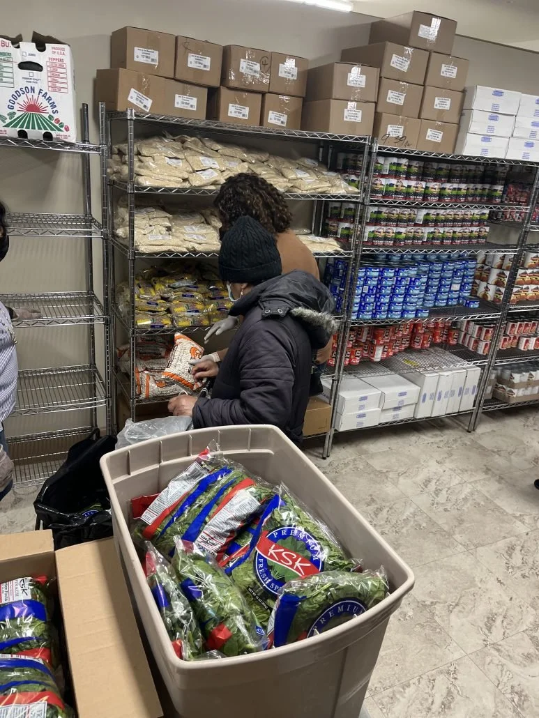 People shopping for groceries in a store aisle, with shelves stocked with packaged foods and a large bin of packaged produce in the foreground.