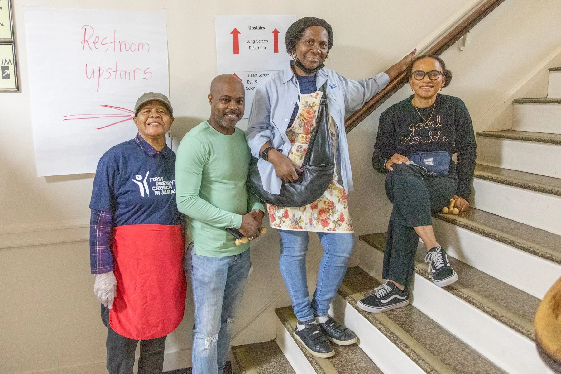 Four people standing on indoor stairs with a whiteboard and signage indicating the upstairs direction in the background.