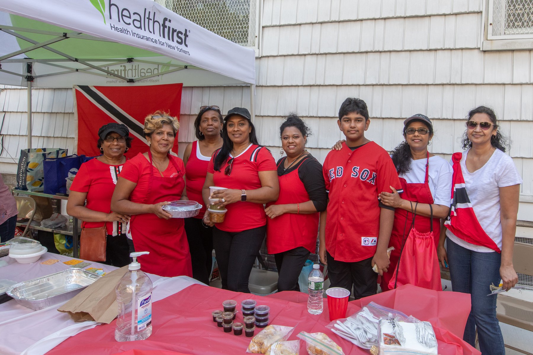 Group of people, mostly women, standing behind a table with drinks and food, at a celebration or gathering, with a banner and a flag in the background.