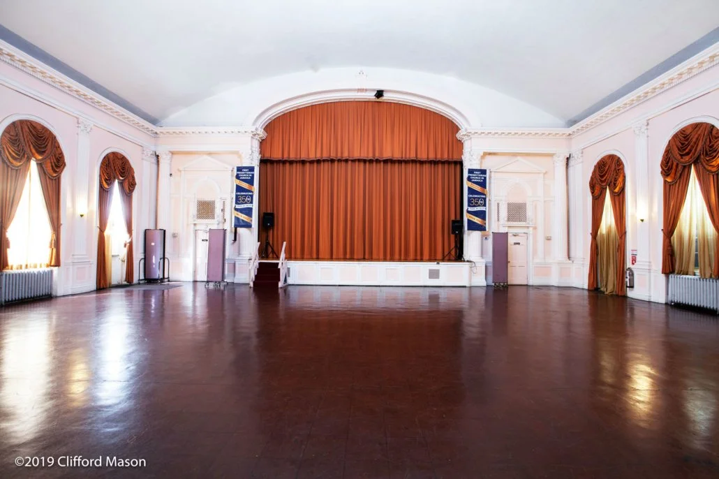 An empty banquet hall with a stage and orange curtains, tall windows with gold curtains, and wood flooring.