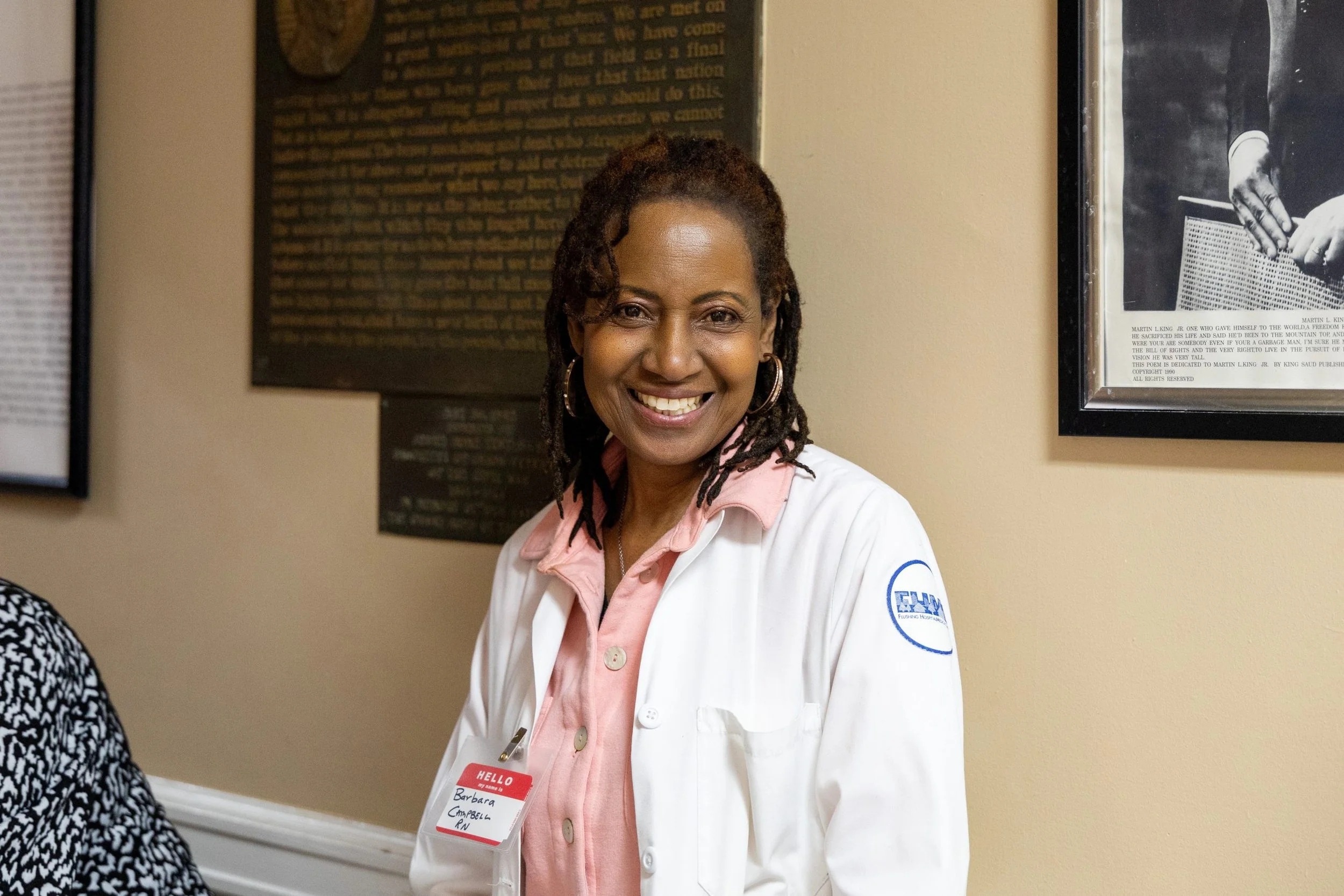 A smiling woman with dark skin and dreadlocks wearing a white coat and pink shirt, standing in front of framed posters on a beige wall.