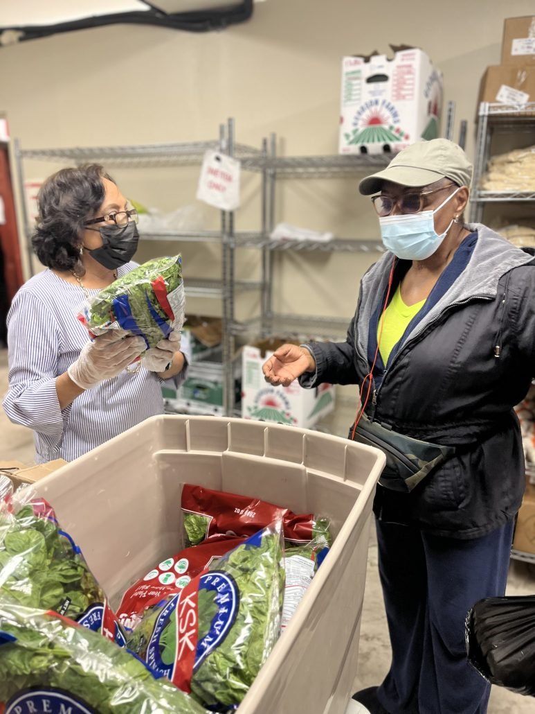 Two women wearing masks and gloves talking near a large container of packaged salad greens inside a storage room or warehouse.