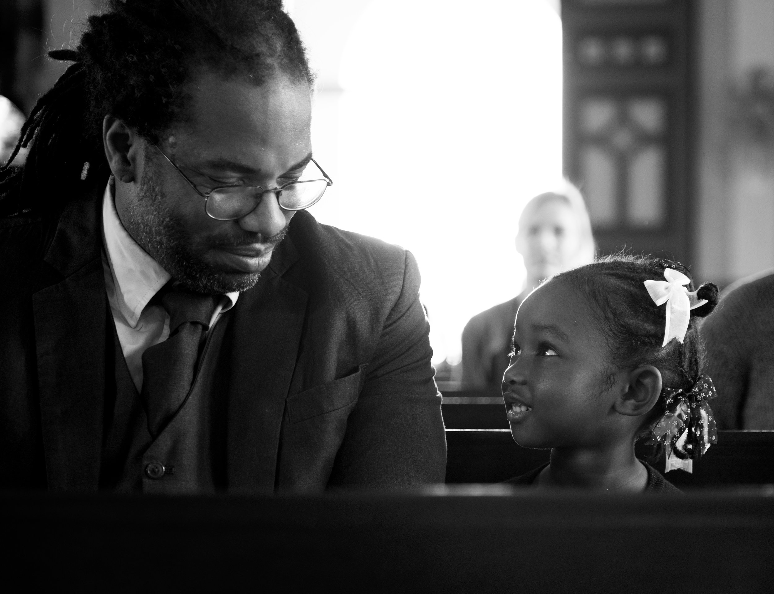 A man and a young girl having a serious conversation in a church, both looking at each other. The man wears glasses and a suit, while the girl has a ribbon in her hair.