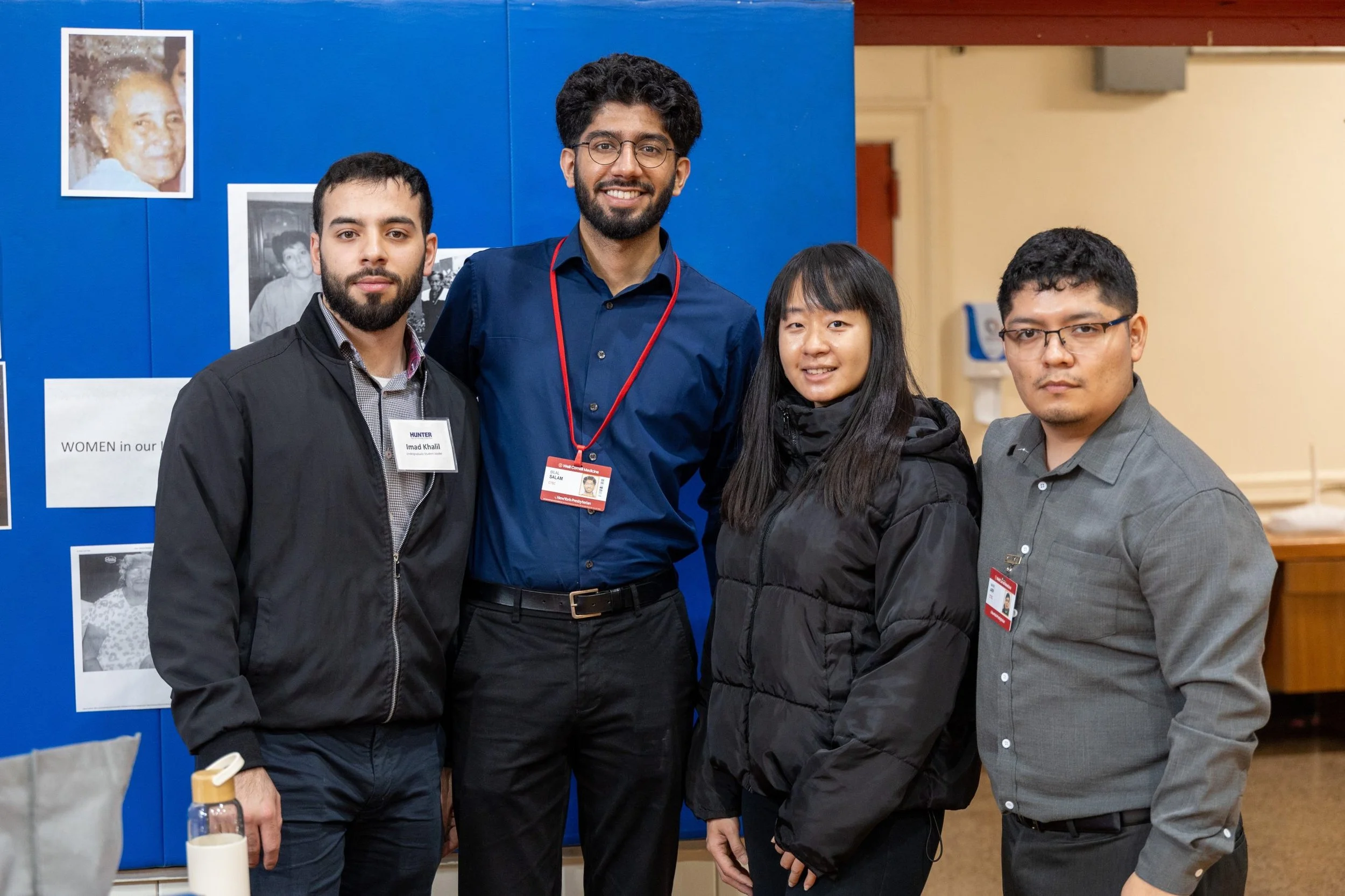 Group of four young adults posing together indoors, two men and two women, at a community event or conference, standing in front of a blue display board with photos and text.