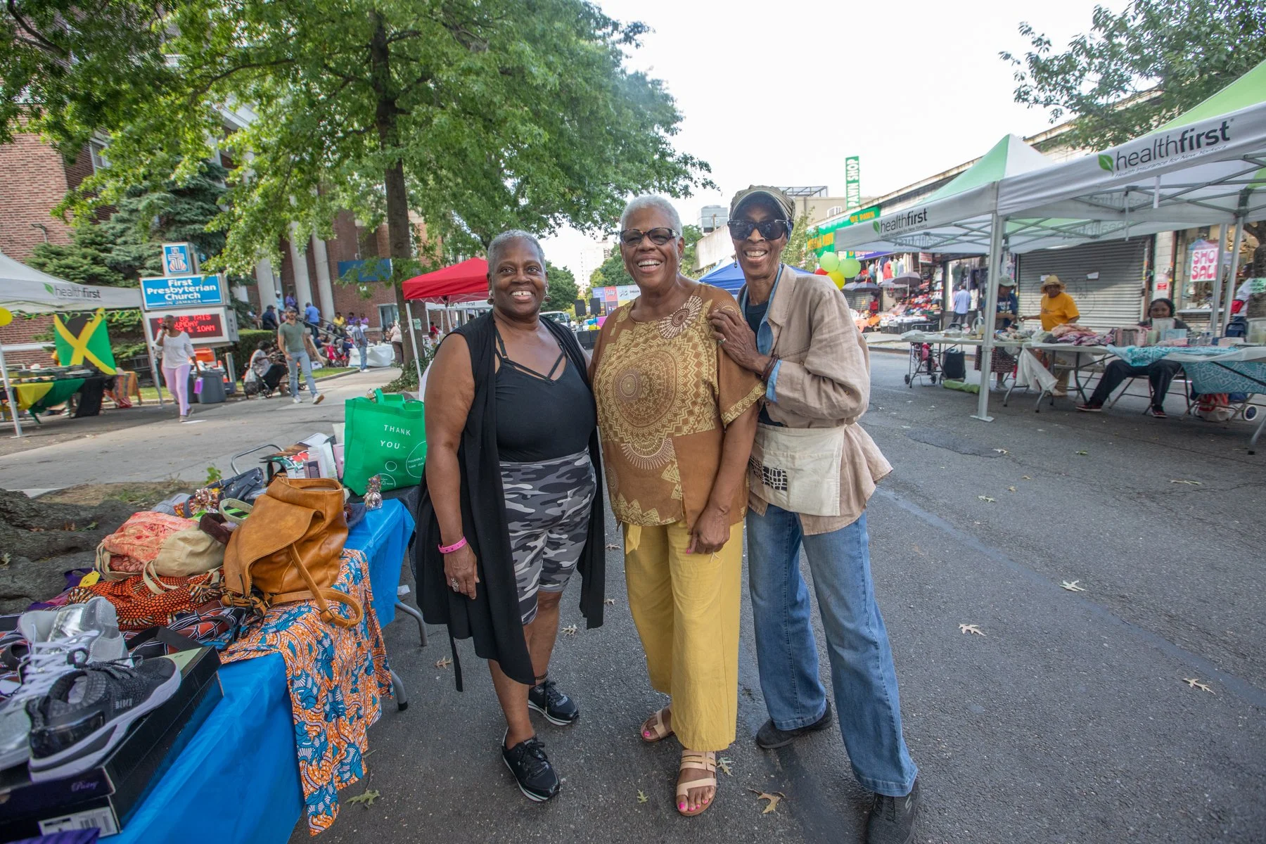Three women standing and smiling at a street market during the day, with tents and vendors in the background, and trees overhead.