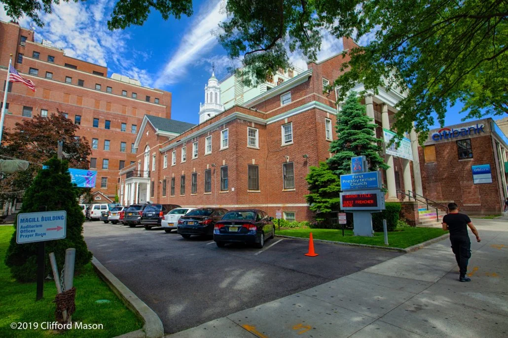 A church building with brick exterior, surrounded by parked cars and trees, with signs pointing to the auditorium, office, and prayer room. A man is walking on the sidewalk, and a digital sign displays a message. The sky is blue with some clouds.