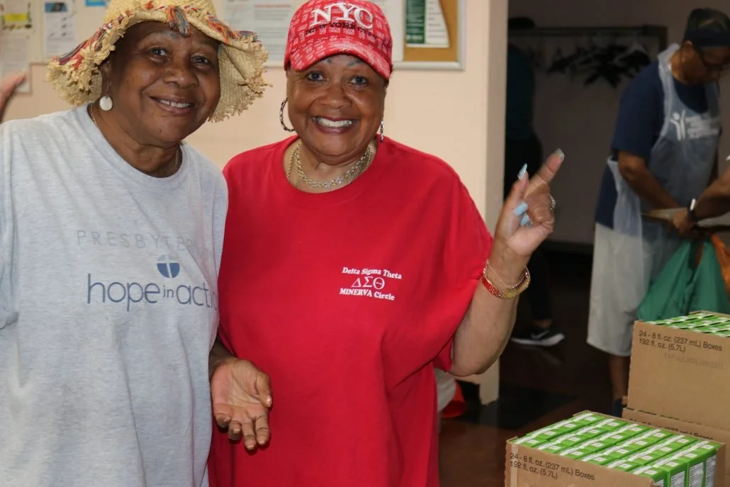 Two smiling women, one wearing a straw hat and a Hope in Action t-shirt, the other wearing a red t-shirt and a red cap that says NYC, standing next to boxes of canned goods, in a community service setting.