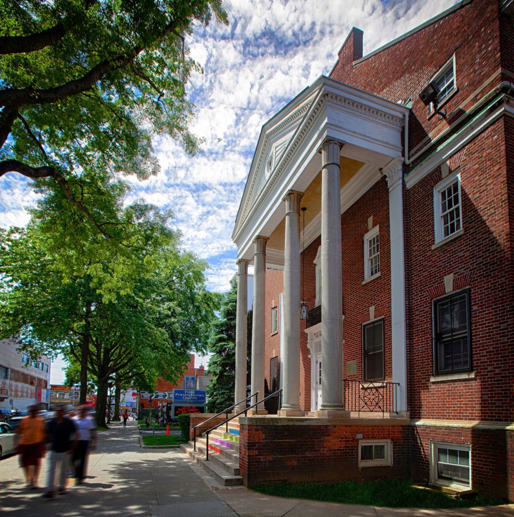 Street scene with a brick building featuring a white front porch with columns, trees lining the sidewalk, and people walking along the street on a partly cloudy day.