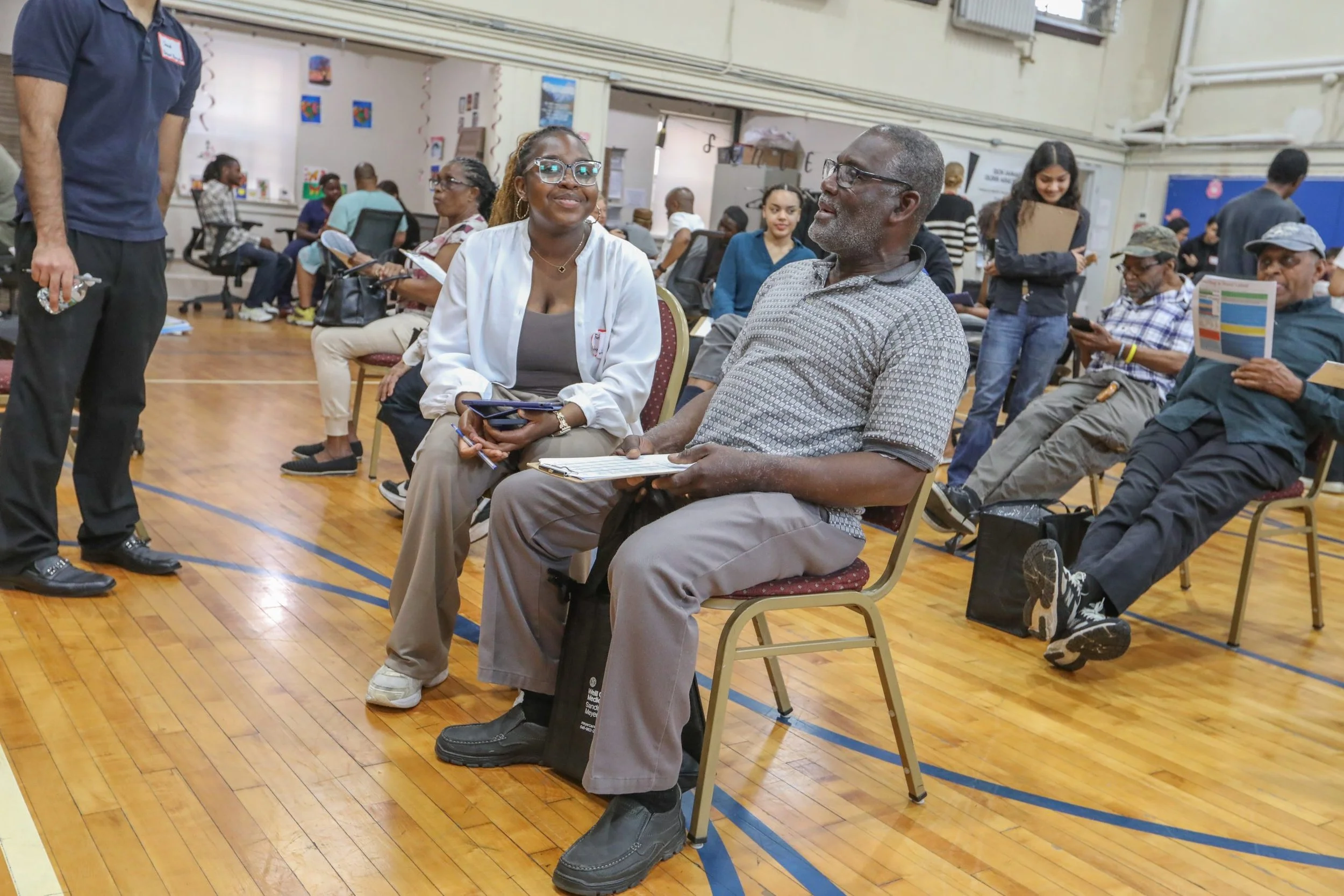 A group of people sitting and talking in a community center or school gymnasium, with some standing in the background. Two people are seated in the foreground, engaged in conversation.
