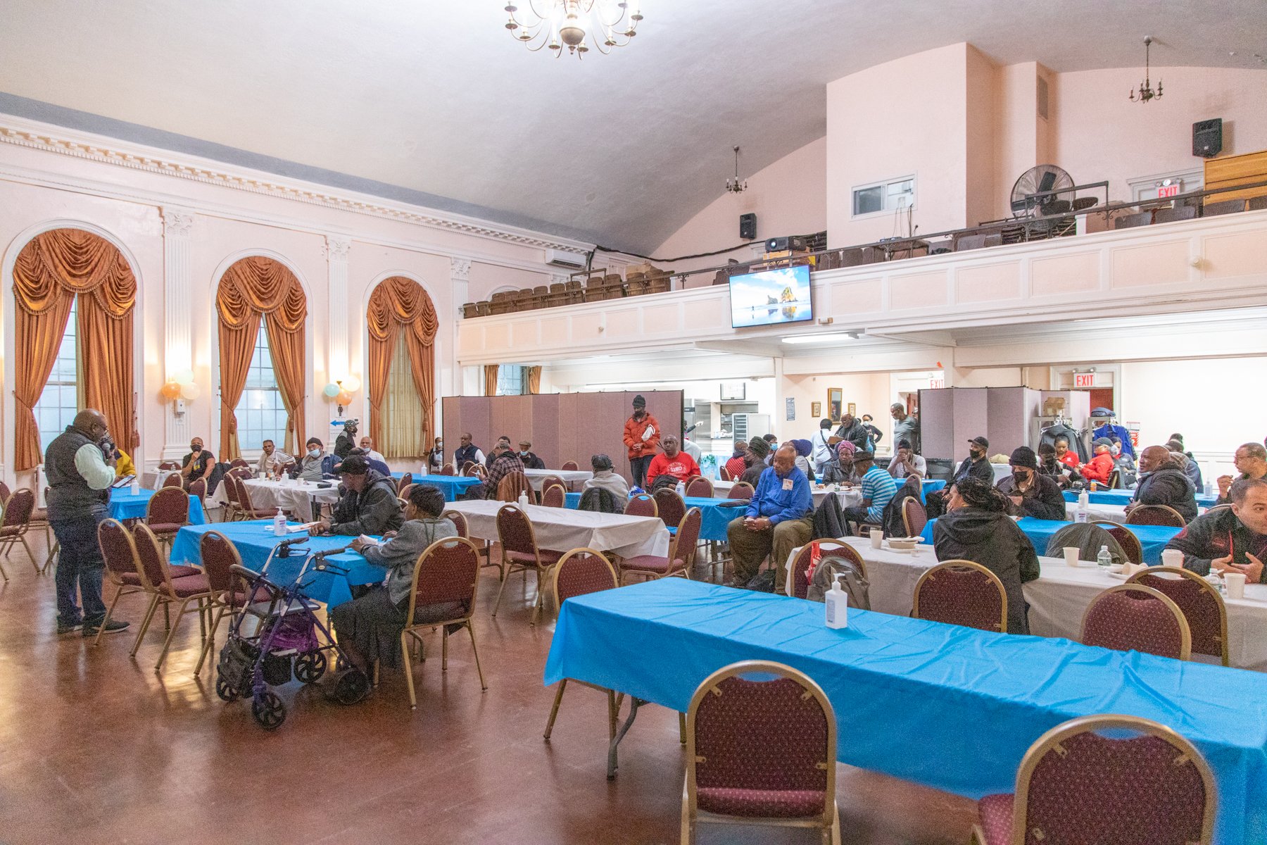Community gathering in a decorated hall with high ceilings, large windows with golden curtains, and a balcony. People seated at tables covered with white and blue cloths, wearing masks, and listening to a speaker at the front.