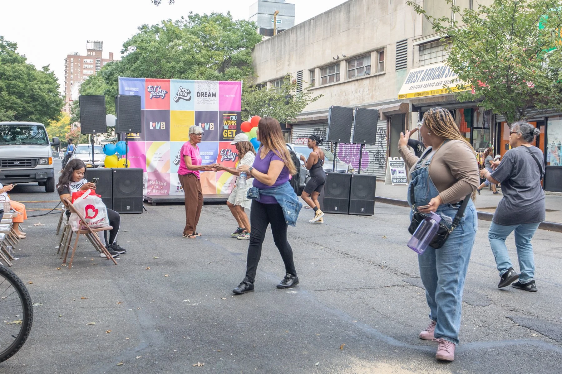 A community outdoor event with a small stage set up against a backdrop featuring colorful squares with motivational words like "Dream," "Love," and "Live." People are dancing and socializing on a paved street, with some sitting, a woman in a pink shi