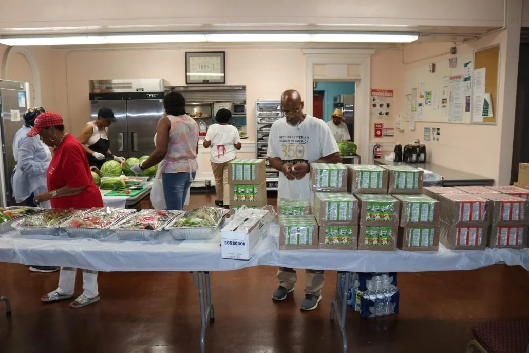 People working together in a kitchen, preparing and packaging food, with shelves, refrigerators, and a bulletin board in the background.