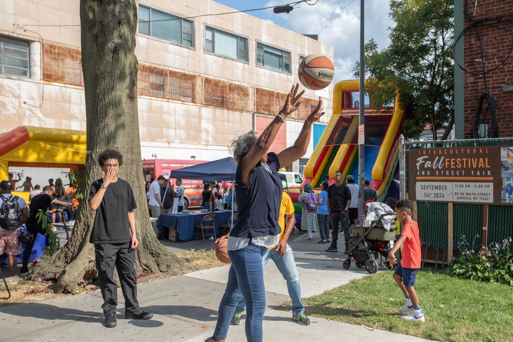 A woman shooting a basketball near a bounce house at an outdoor fall festival event with people and tents in the background.
