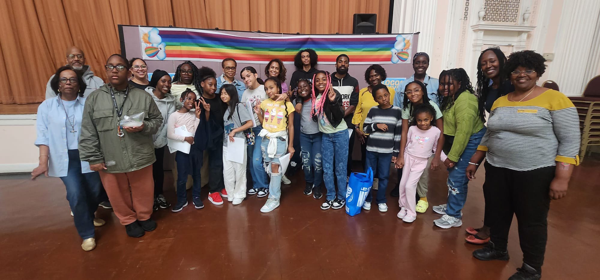 Group of children and adults posing for a photo in an indoor event space with a wooden wall and a rainbow banner in the background.