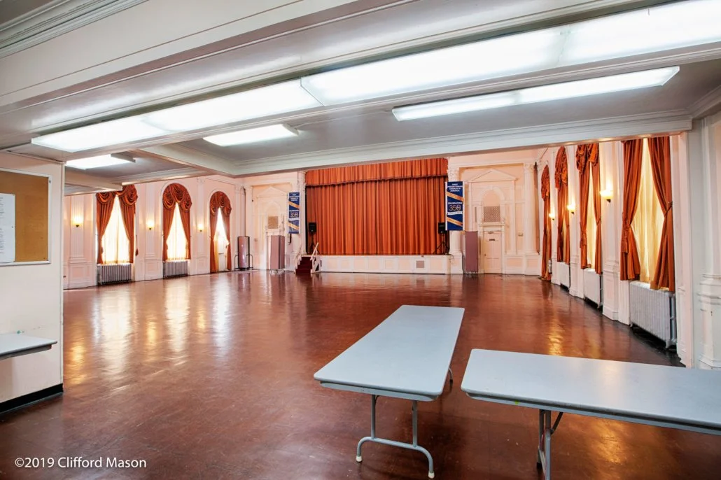 Empty event hall with a stage, orange curtains, and folding tables in the foreground, illuminated by natural light through windows with orange drapes.