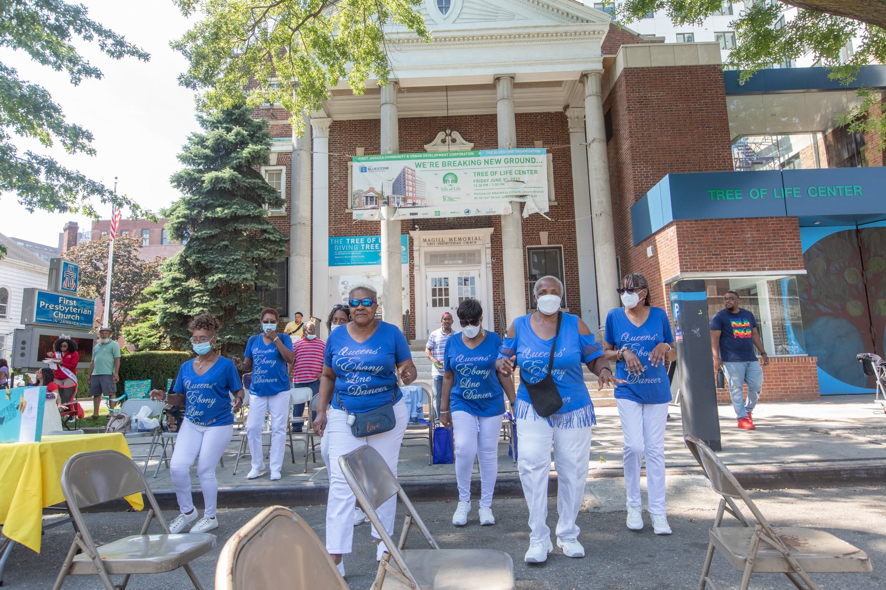 Group of women wearing matching blue t-shirts and white pants, walking in front of a brick church building with a sign that reads "First Presbyterian Church in Jamaica," and a banner that says "We're breaking new ground... Tree of Life Center," durin