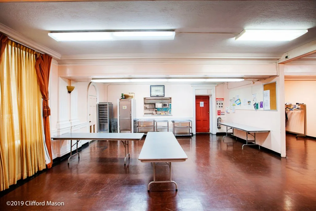 Empty dining or event room with tables, a fridge, bulletin board, and a red door, with yellow curtains on the left side.