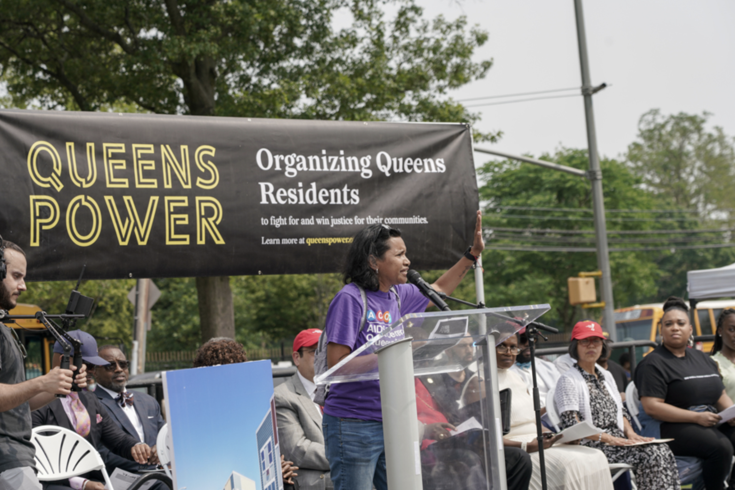 A woman speaking at a podium during a protest or rally, with a large banner behind her that reads 'Queens Power, Organizing Queens Residents to fight for and win justice for their communities.' Several other people are seated on stage, some wearing hats, and one person is holding a video camera. Trees and power lines are visible in the background.