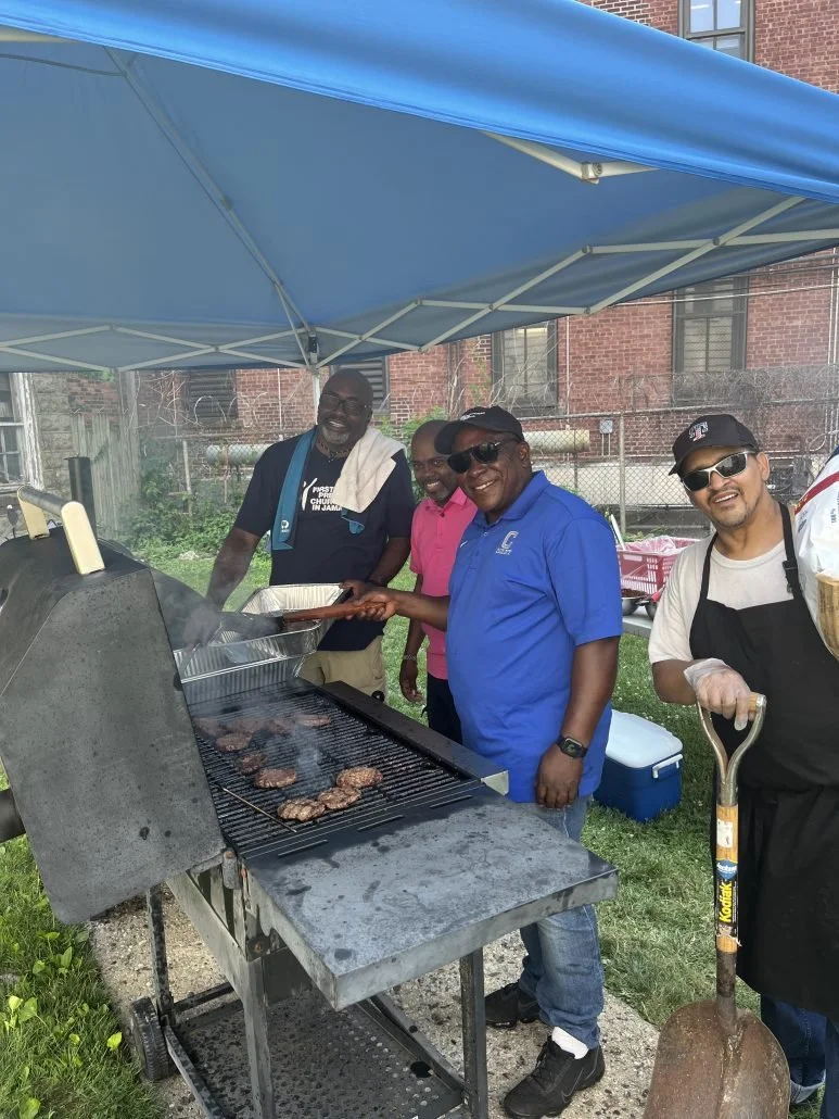 Four men standing next to a barbecue grill under a blue canopy, cooking burgers outdoors. One man is wearing a black apron and gloves, holding a spatula, while the others are smiling and wearing casual shirts.