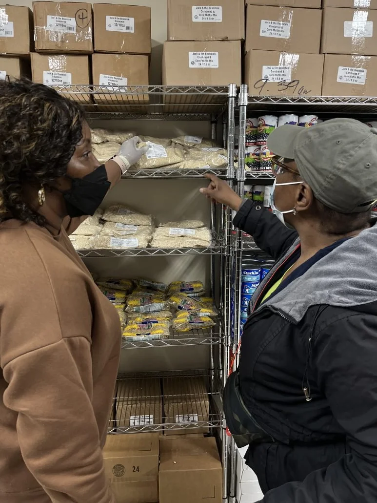 Two women in masks and jackets stand in front of metal shelves filled with bulk food items in a storage or warehouse setting. One woman points at the shelves while the other reaches for a package.