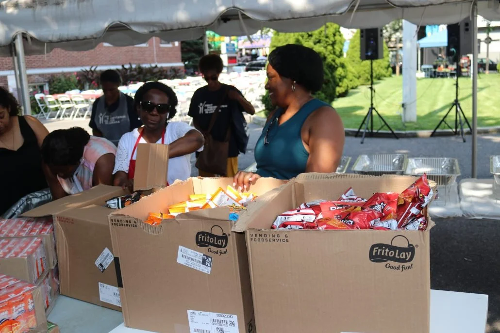 People at an outdoor event under a tent, sorting and distributing snacks like chips from large cardboard boxes, with a grassy area, trees, and speakers in the background.