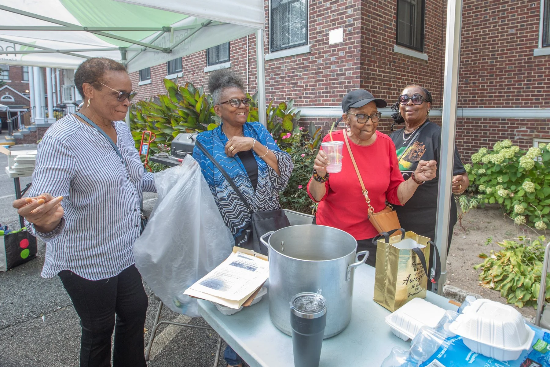 Four women standing behind a table at an outdoor event, with one woman holding a drink, in front of a brick building and plants.