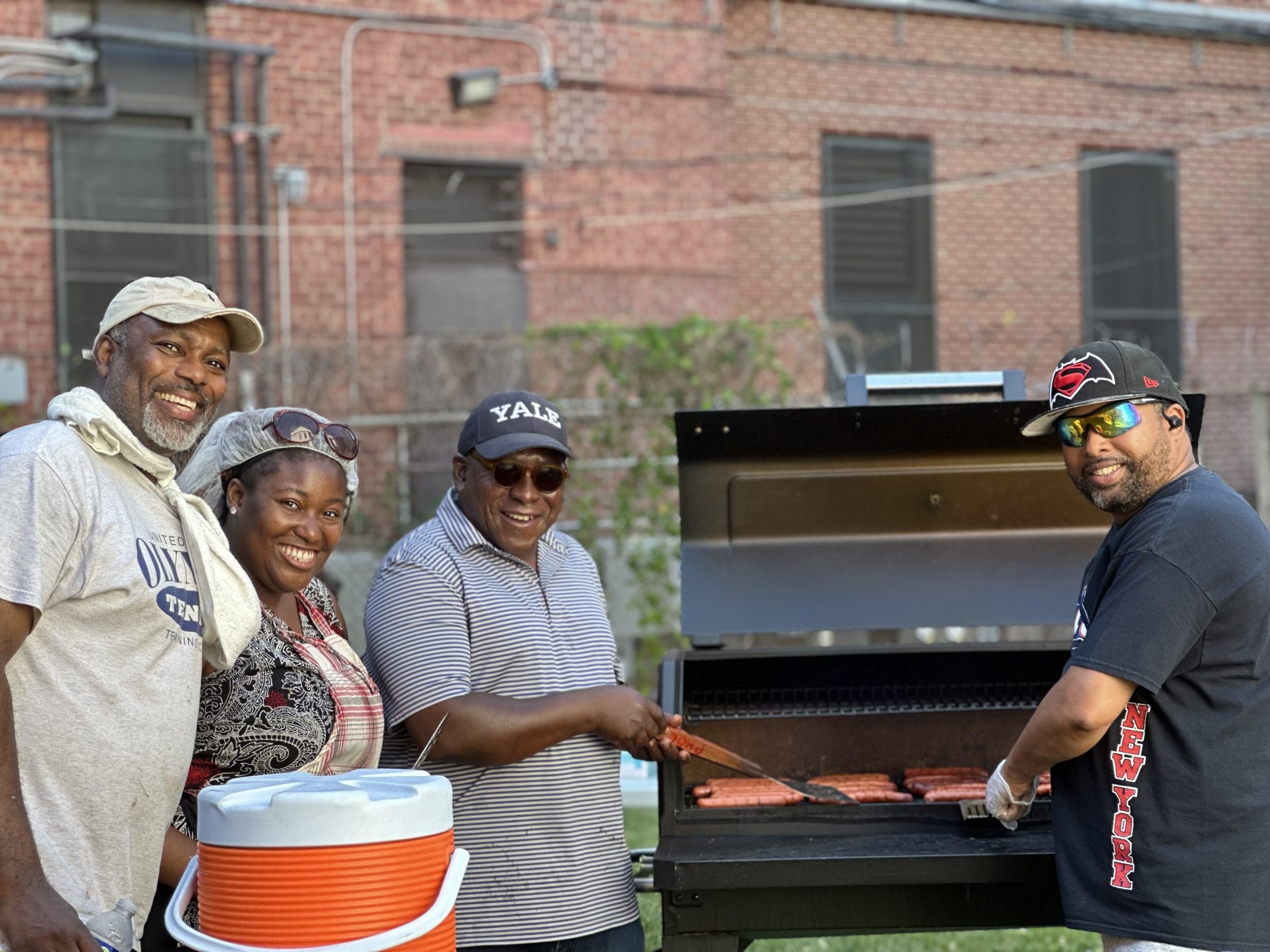 Five people, three men and two women, are gathered around a barbecue grill outdoors, cooking and smiling. One man is flipping sausages on the grill, and the group appears to be enjoying a barbecue gathering.