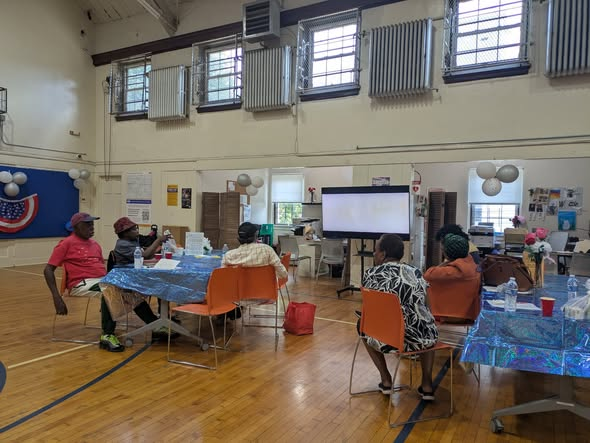 Group of people sitting at tables in a decorated community room, with patriotic decorations like balloons, banners, and a large screen at the front.