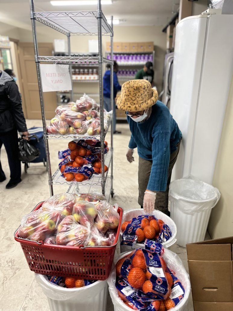A woman in a blue sweater, leopard print hat, and face mask is stacking plastic bags of oranges in a store. There are plastic bags of apples in a red basket and hanging on a shelf nearby, with other shoppers in the background.