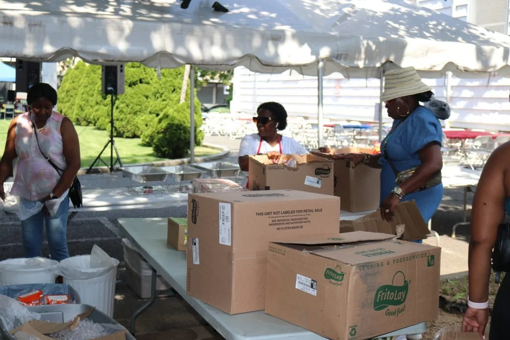 People working at a food service booth under a white canopy, with boxes of supplies on the table, outdoors with a building and greenery in the background.