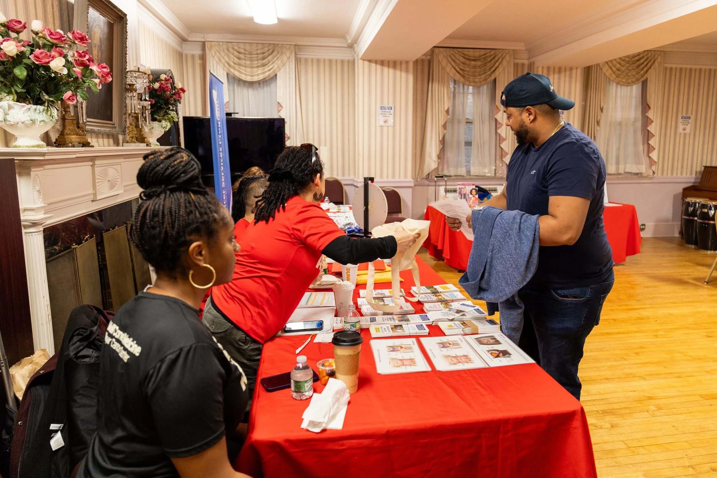 People at a table with informational brochures and registration materials, interacting with a man at an indoor event.