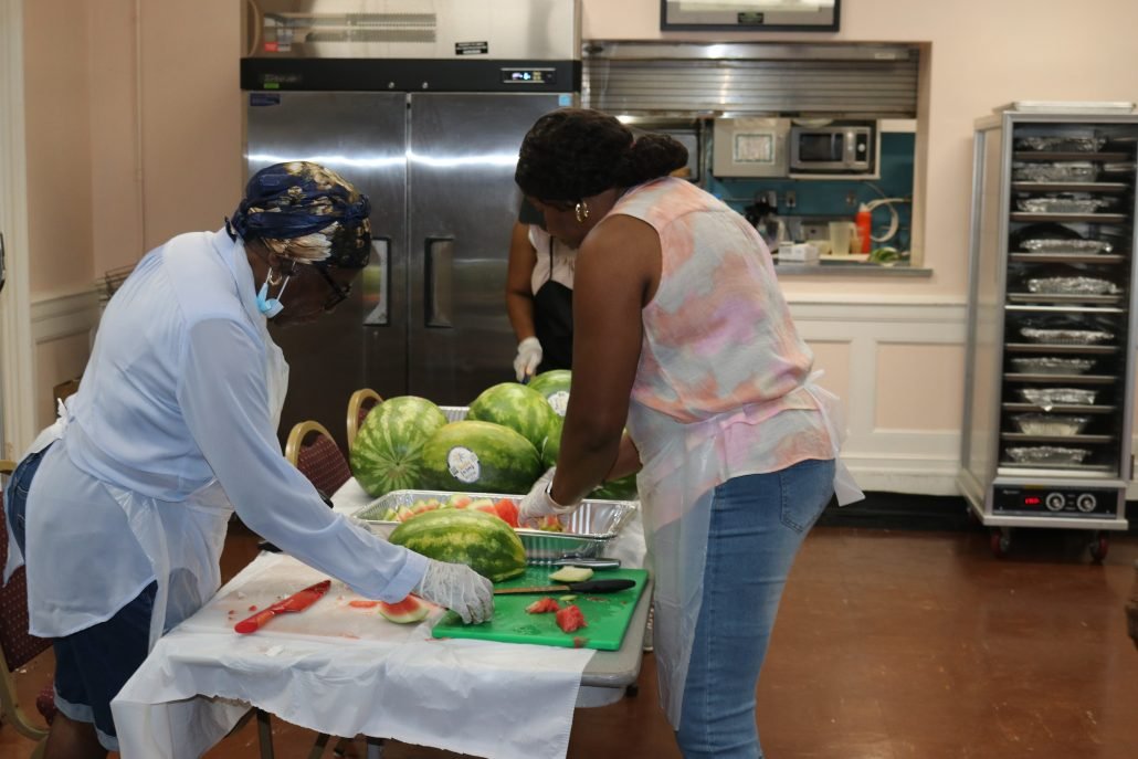 Two women are slicing watermelon on a table with additional watermelons and a knife. They are in a kitchen or food preparation area.