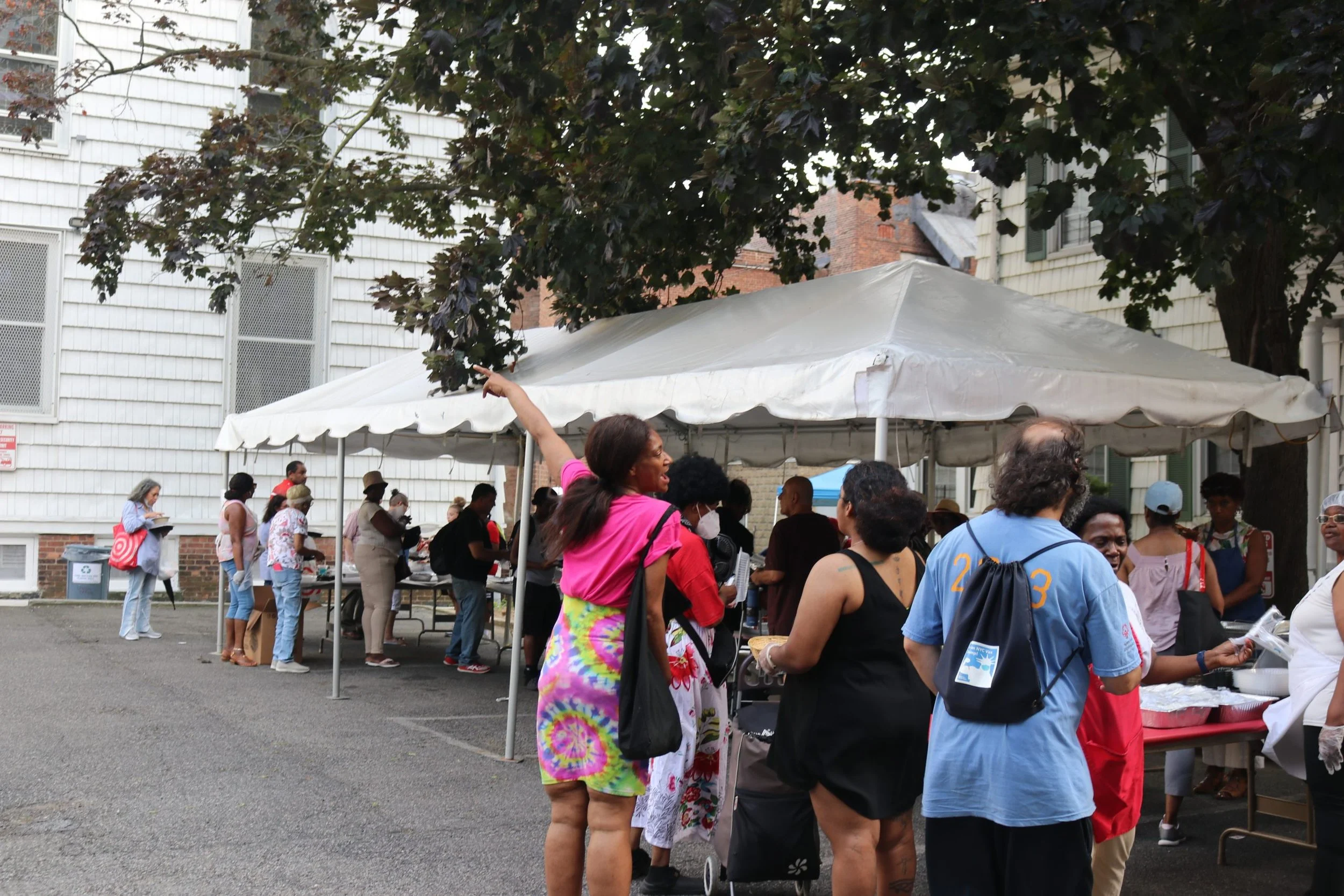 People standing in line and gathering around under a white canopy tent outside of a building, engaging in a community event or festival.