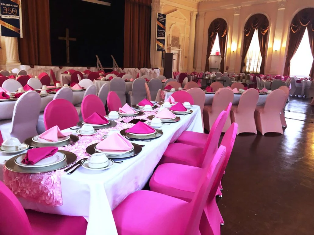 A banquet hall decorated with tables and chairs in shades of pink, with pink napkins and place settings, gold chargers, and a floral table runner. The room has tall windows with dark curtains, and a stage at the front with a cross backdrop.