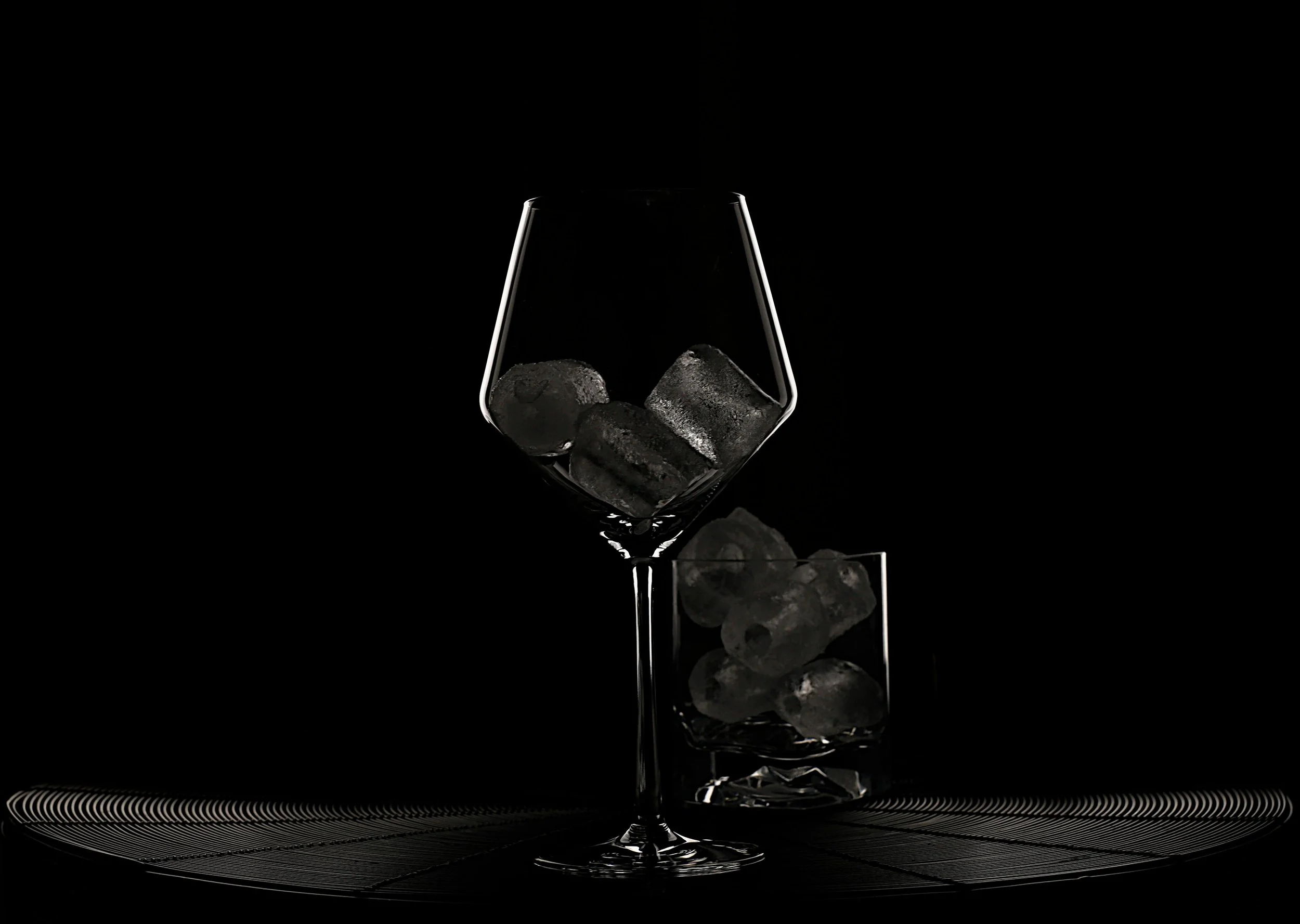 A glass with ice cubes balanced inside and a second glass filled with ice cubes beneath it, set against a dark background.
